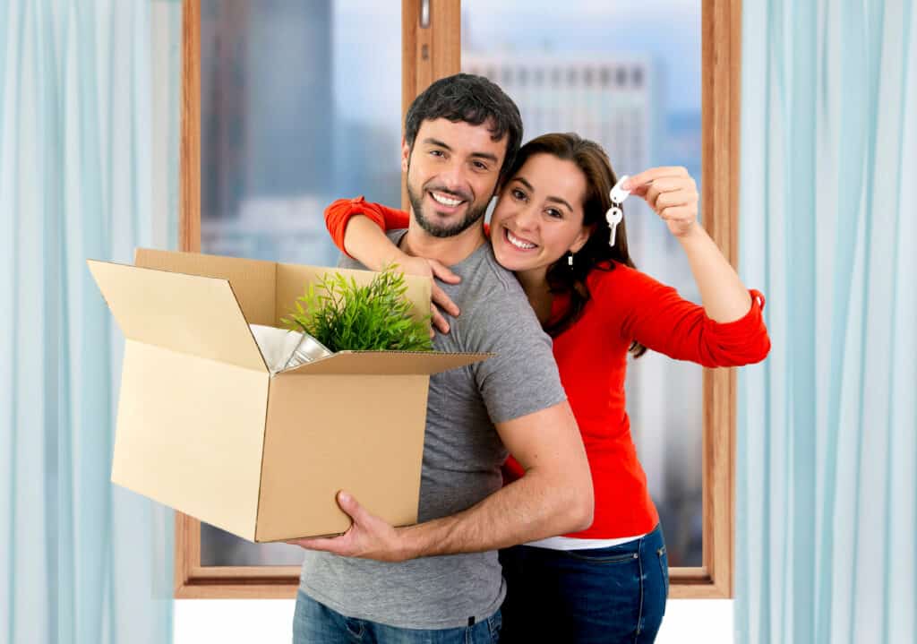 young happy Hispanic couple moving together in a new flat or apartment carrying cardboard boxes home belongings holding house key smiling in housing and real state concept